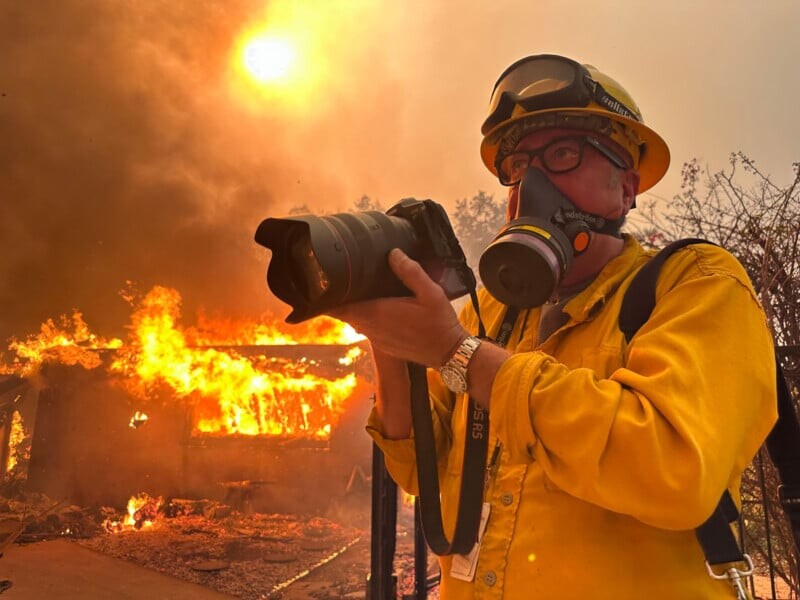 A person in a yellow fire-resistant suit and helmet, wearing a respirator, holds a camera with a burning building and a smoky, red-tinted sky in the background. The sun appears hazy due to the smoke.