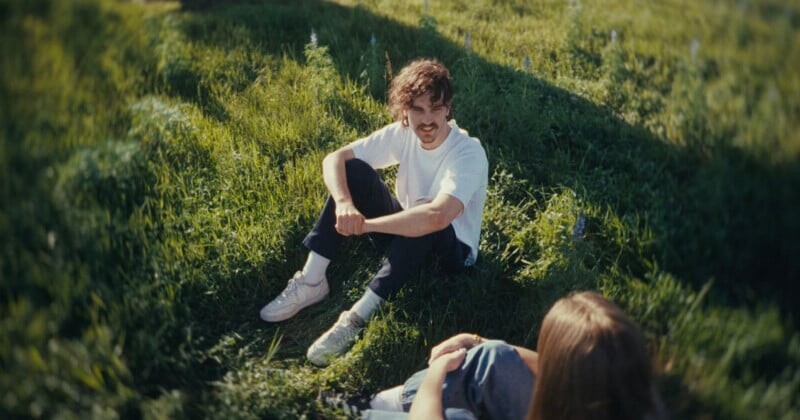 A man in a white t-shirt and dark pants sits on grass, smiling and talking with a person whose back is to the camera. Sunlight casts shadows, and the setting appears calm and natural.
