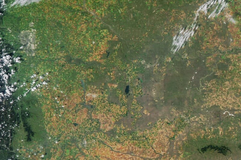 Satellite image showing a patchwork of green and brown agricultural fields, scattered clouds, and a mix of forested and open land, with mountains visible in the lower left corner.