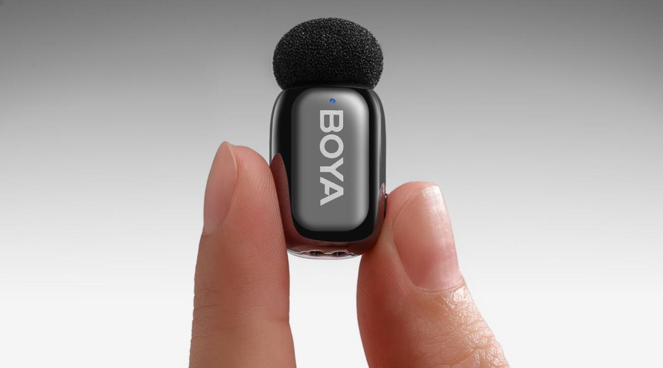 A close-up of two fingers holding a small black BOYA-branded wireless microphone with a foam windscreen against a plain gradient background.