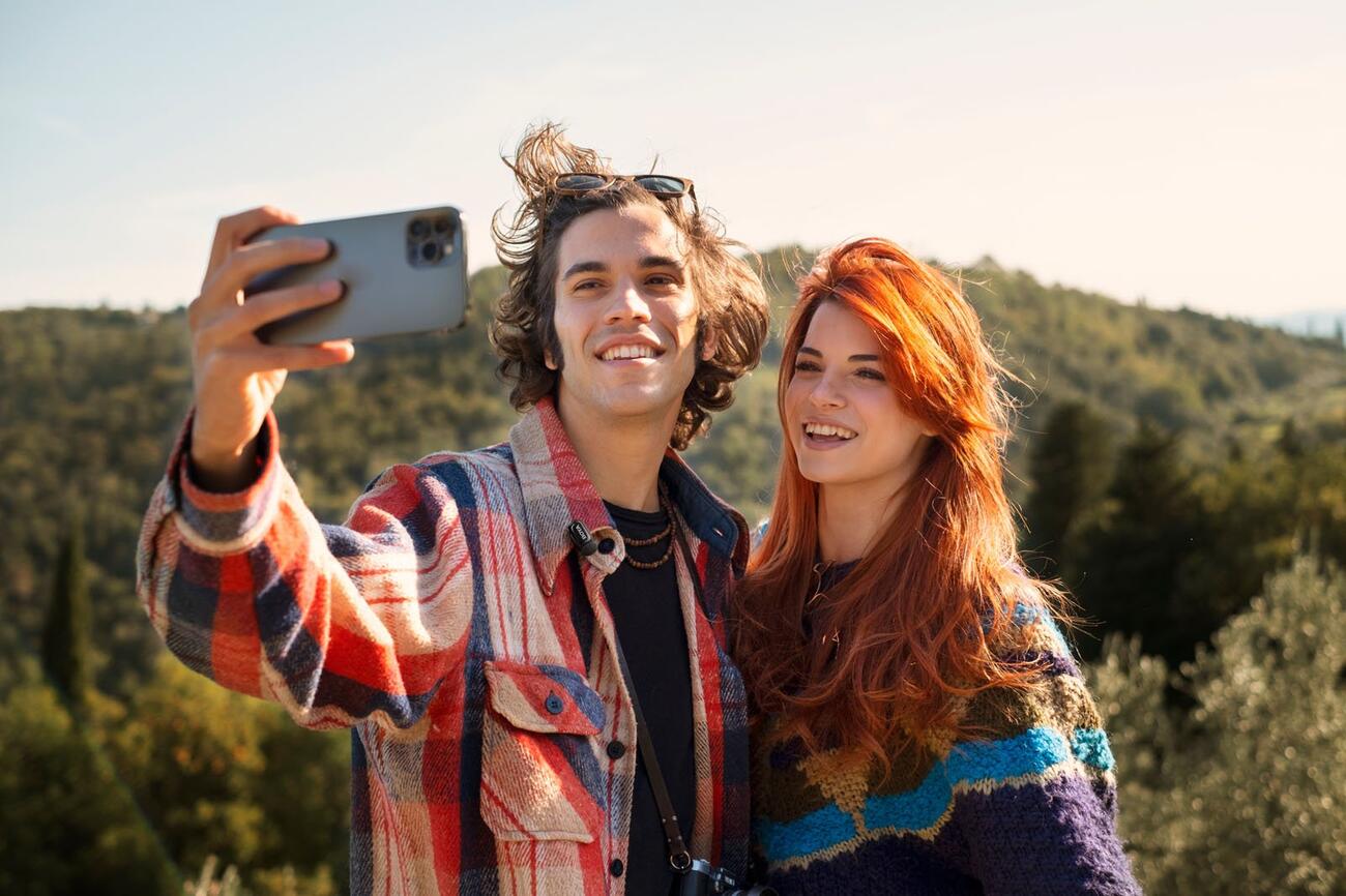 Two young adults smiling and taking a selfie outdoors, with green hills and trees in the background on a sunny day.