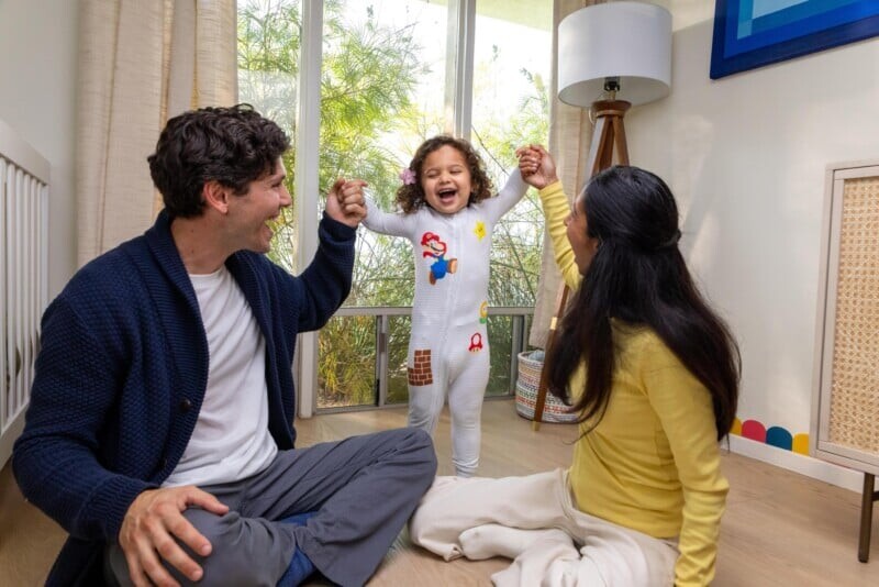 A smiling child in pajamas stands holding hands with two adults, who are seated on the floor in a bright room with large windows and cheerful decor.