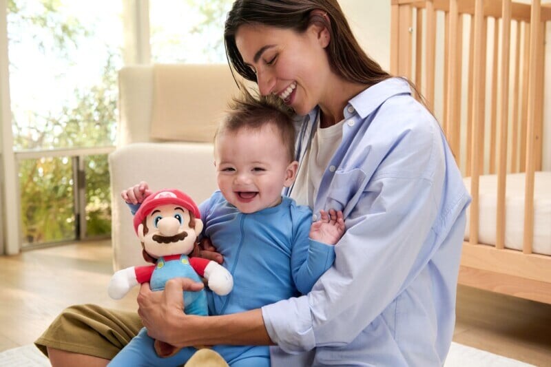 A smiling woman sits on the floor holding a happy baby dressed in blue pajamas, who is holding a plush Mario toy, in a bright, cozy nursery with a crib in the background.