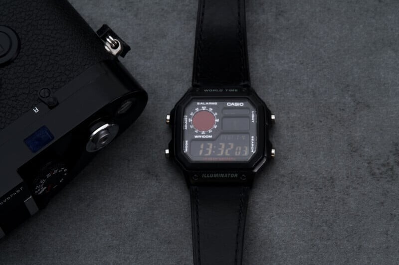 A Casio digital wristwatch with a black leather strap sits on a gray surface next to a vintage film camera. The watch displays the time, date, and world time features on its screen.