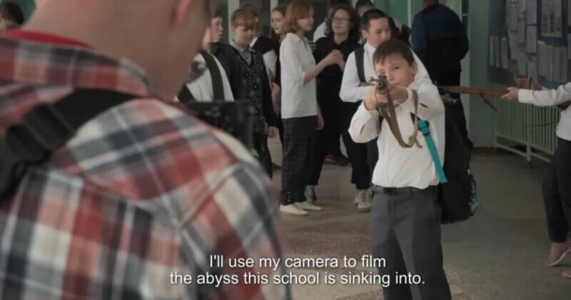 A boy in a school uniform points a rifle at a man with a camera, while other students watch in the background.