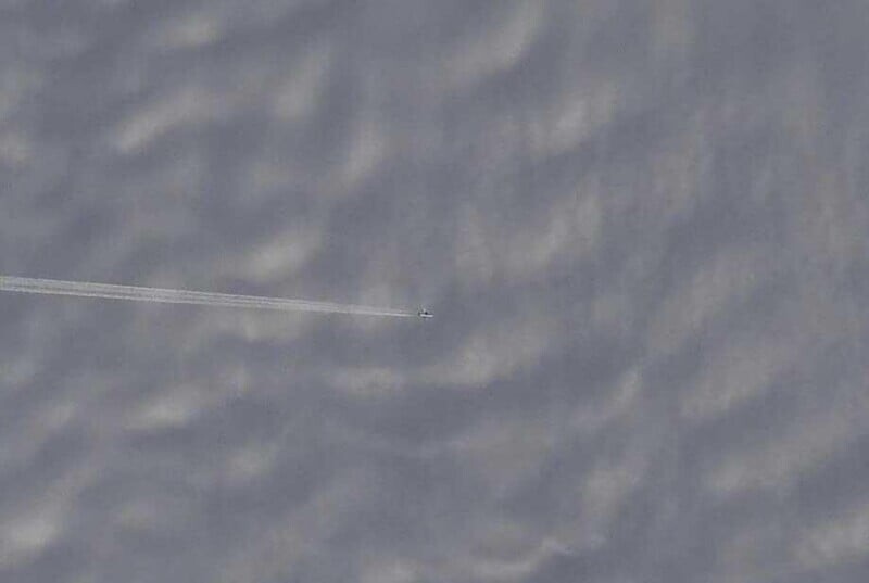 Далёкий самолет выпускает тонкий след, пролетая над облачным небом, выглядящим маленьким на фоне текстурованных облаков нижнего уровня.