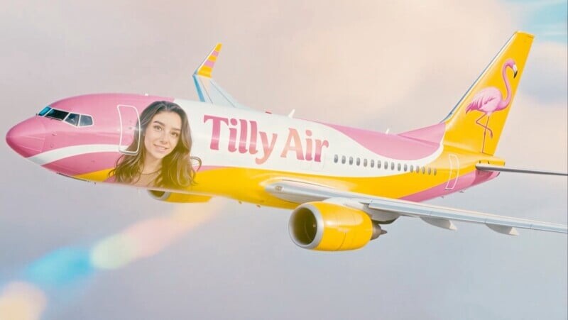Розовый и жёлтый самолёт с надписью 'Tilly Air', большой фотографией лица молодой женщины и графикой фламинго на хвосте, летит на фоне облачного неба.