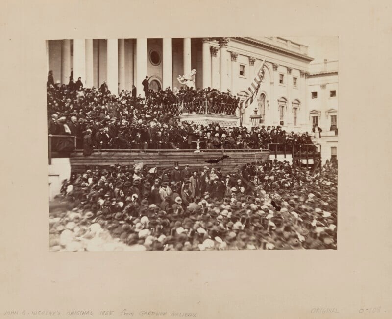 A large crowd gathers outside a government building for an event, with people standing on steps and balconies. An American flag hangs nearby, and a speaker addresses the audience from a platform.