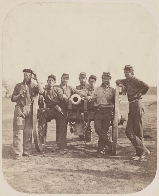 Seven Civil War soldiers in uniform pose around a large cannon on a grassy field, with several tents visible in the background.