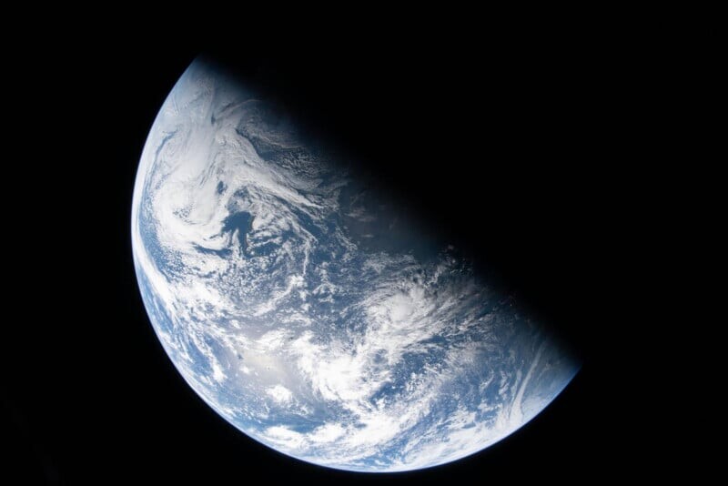A view of Earth from space, showing swirling white clouds over blue oceans and land, with nearly half the planet in shadow against the blackness of space.