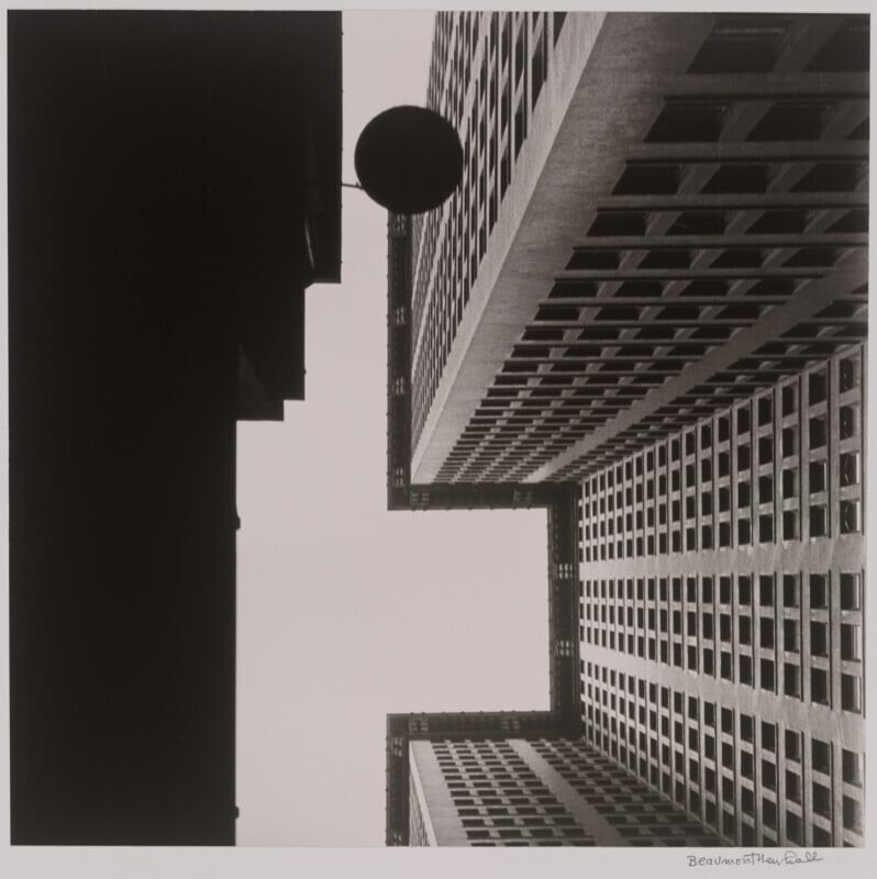 Black and white photo looking up at tall, geometric buildings with many windows, forming sharp angles against the sky; a circular object is suspended between the buildings.