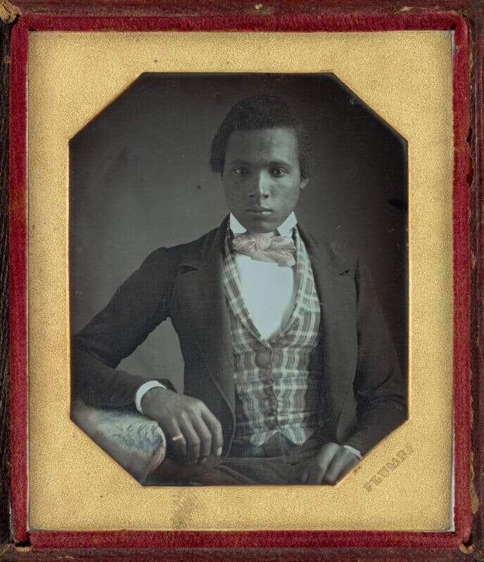 A daguerreotype portrait of a young Black man wearing a dark coat, a plaid vest, and a light-colored bow tie.